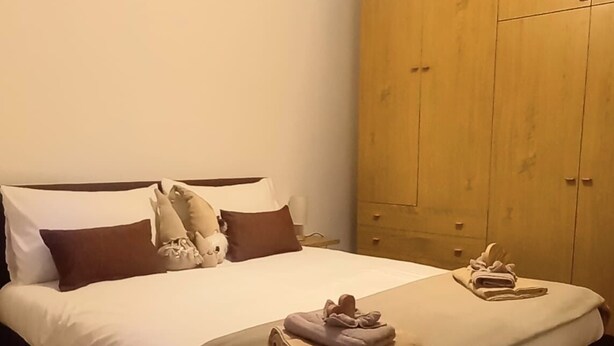 Uma cama bem arrumada com manta e travesseiros marrons, um guarda-roupa de madeira e uma pequena mesa com toalha e livro.