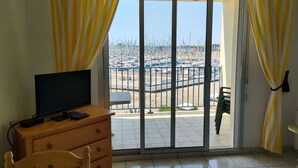 A room with a TV on a wooden dresser, a chair, and a table. It has a sliding glass door leading to a marina with boats. - Appartement 2 pièces sur le Port du Crouesty, à louer pour les vacances (ARZON)