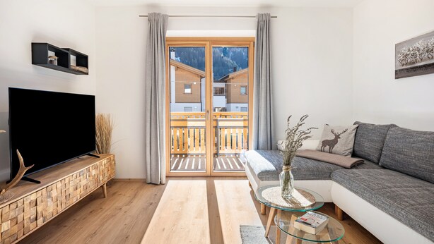 A modern living room with a flat-screen TV, a sofa, a glass coffee table, and a view of a mountain through a sliding door. - Apartment 'Jan's  Am See' with Lake View, Balcony and Wi-Fi (Pracupola)