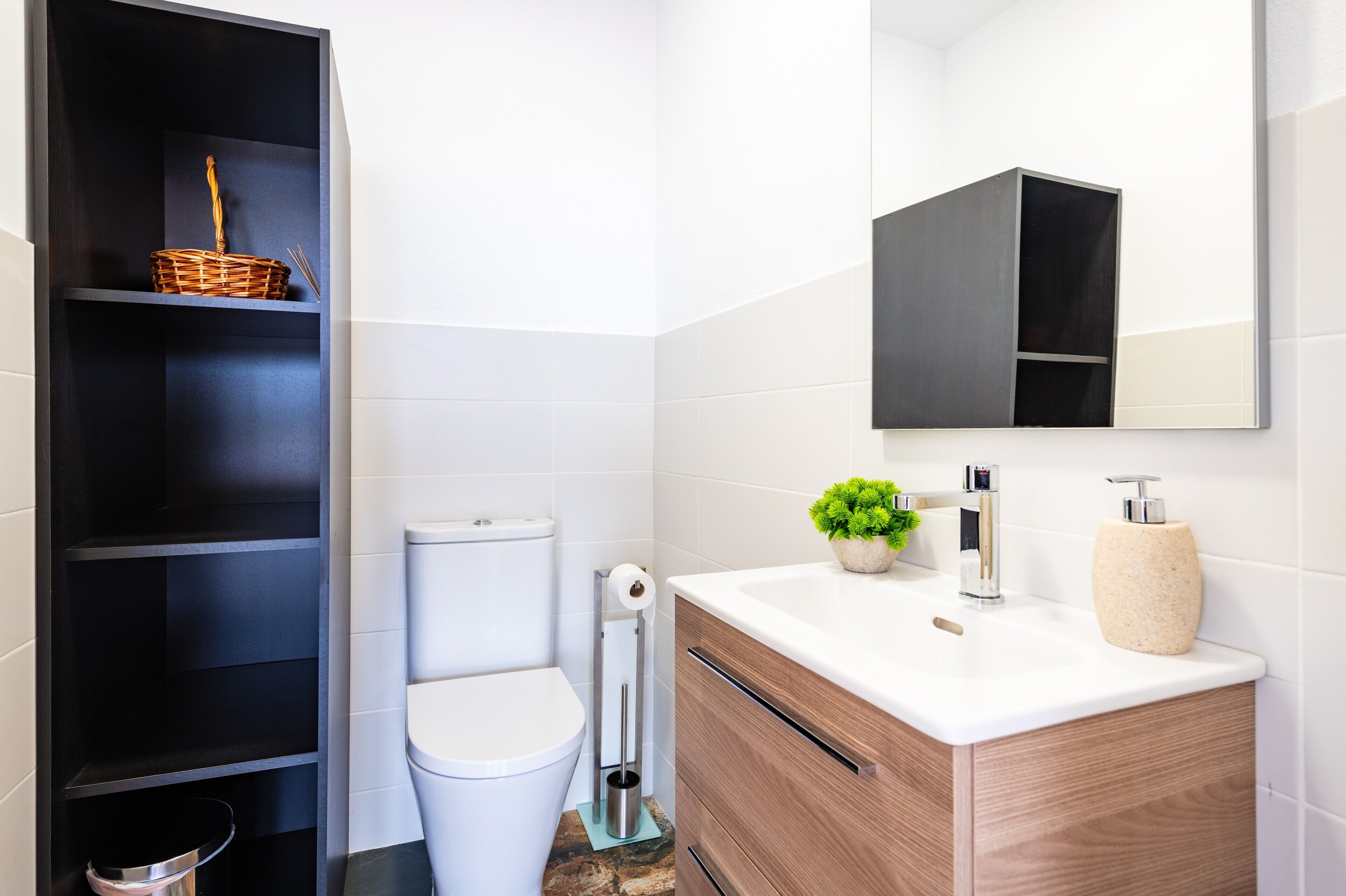 Un baño moderno con inodoro, lavamanos con espejo, estante con cesta y planta.