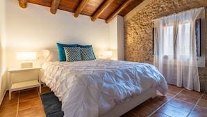 Une chambre avec un mur en pierre, des poutres apparentes en bois au plafond, un lit avec une literie blanche, une table de chevet et une fenêtre ornée de rideaux légers.