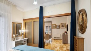 A hotel room with a bed, a desk with a lamp, a dresser, and a large mirror. There is a door leading to another room.
