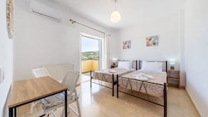 Une chambre avec deux lits, une table, une chaise, un radiateur, un appareil de climatisation, un luminaire au plafond et une vue sur l’extérieur.