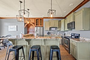 A kitchen with green cabinets, a stainless steel refrigerator, a microwave, and a stove. There are two bar stools and a large island with a marble countertop.