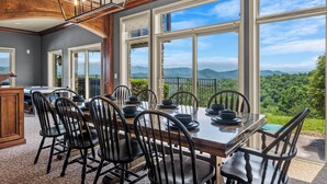 Comedor con una gran mesa dispuesta para seis personas, rodeada de sillas, y vistas a montañas a través de amplios ventanales.