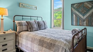 Un dormitorio con cama, mesita de noche, lámpara, ventana con vistas a árboles y decoración en la pared.