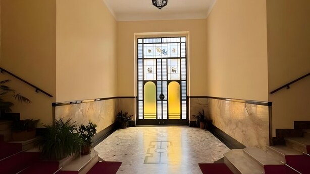 A grand staircase with red carpet runners leading to a large, ornate door with stained glass panels.