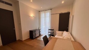 A hotel room with a wooden floor, a bed with white bedding, a small table, a chair, a cabinet with a 'DO NOT PARK' sign, and a window with curtains.
