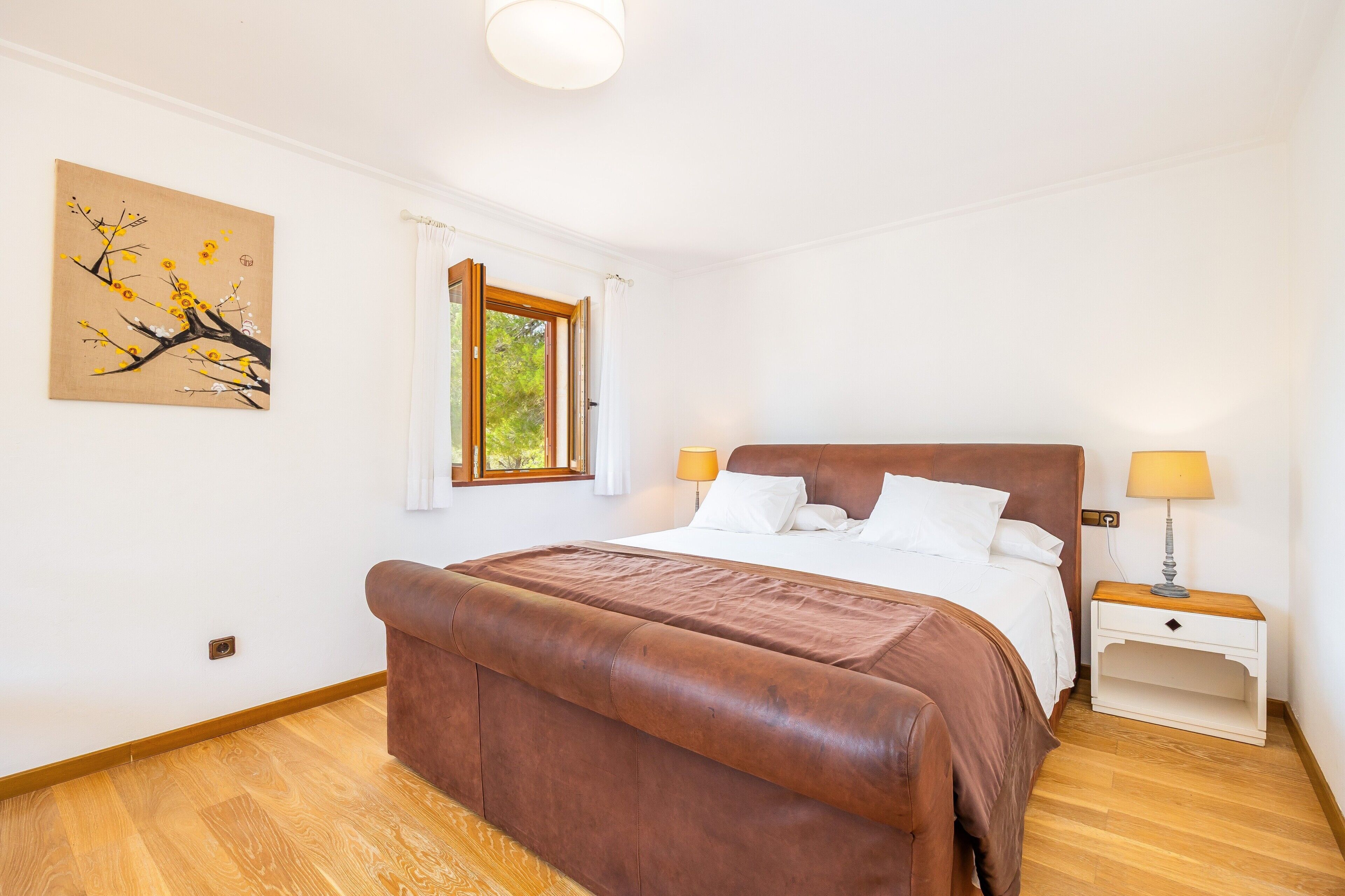A bedroom with a large bed, a nightstand with a lamp, a window with curtains, and a wall painting.