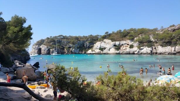 Una playa con aguas turquesas cristalinas, acantilados rocosos y un velero.