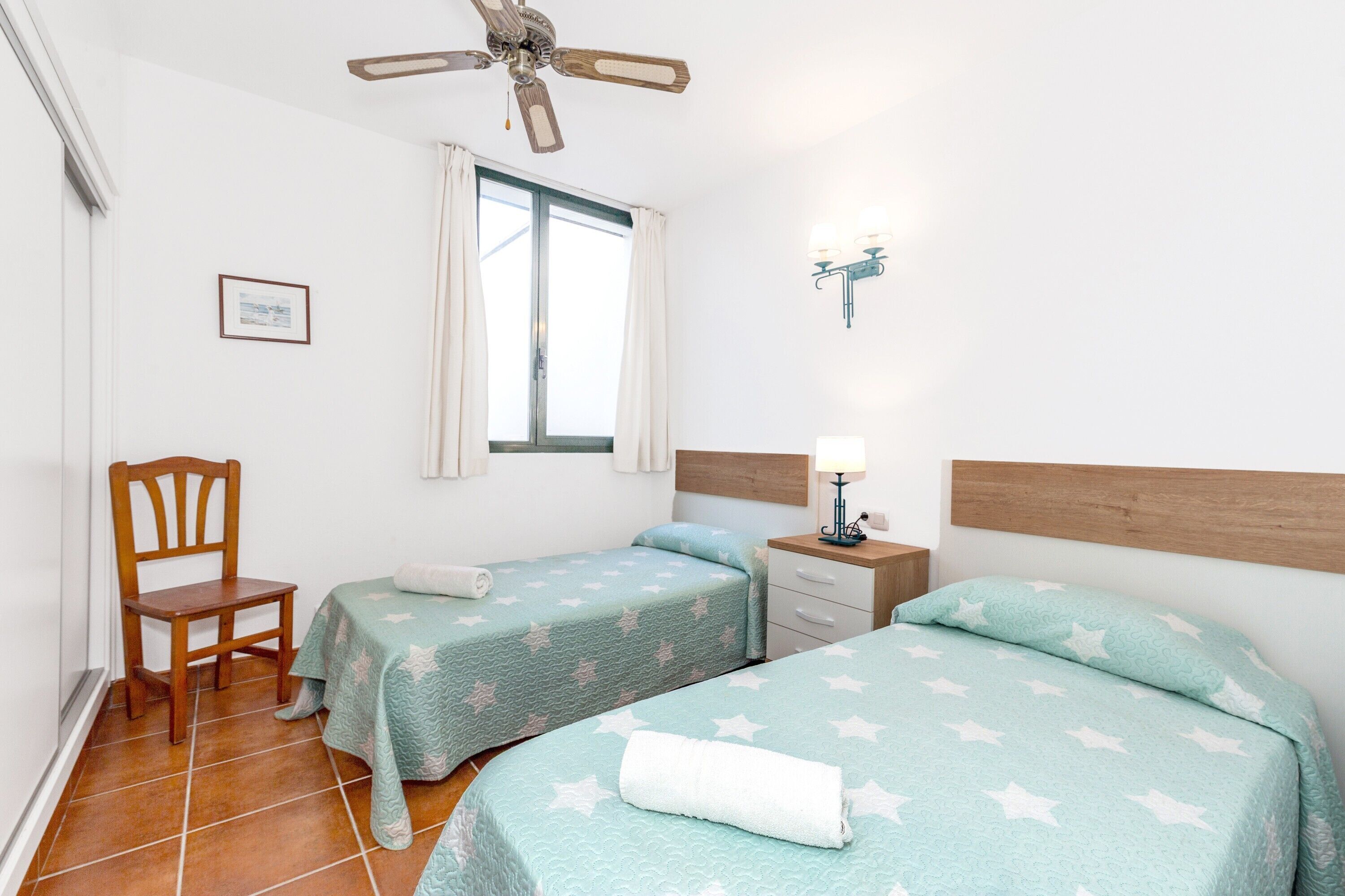 A room with two beds, a chair, a window with curtains, a ceiling fan, a wall-mounted lamp, and a framed certificate on the wall.