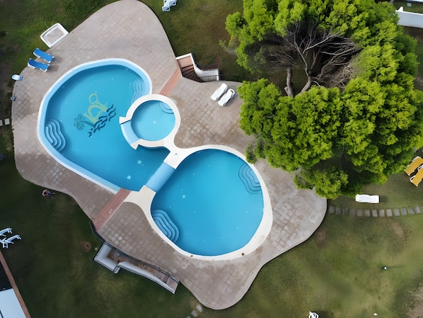 A large swimming pool with multiple sections, surrounded by lounge chairs and a tree.