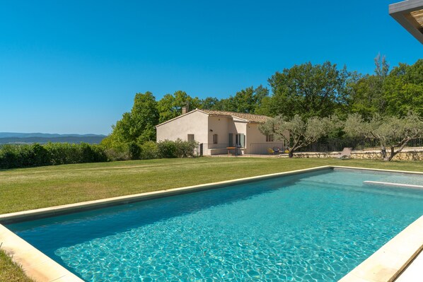 A rectangular swimming pool with clear blue water, surrounded by a well-maintained lawn, a house with a tiled roof, and trees in the background.