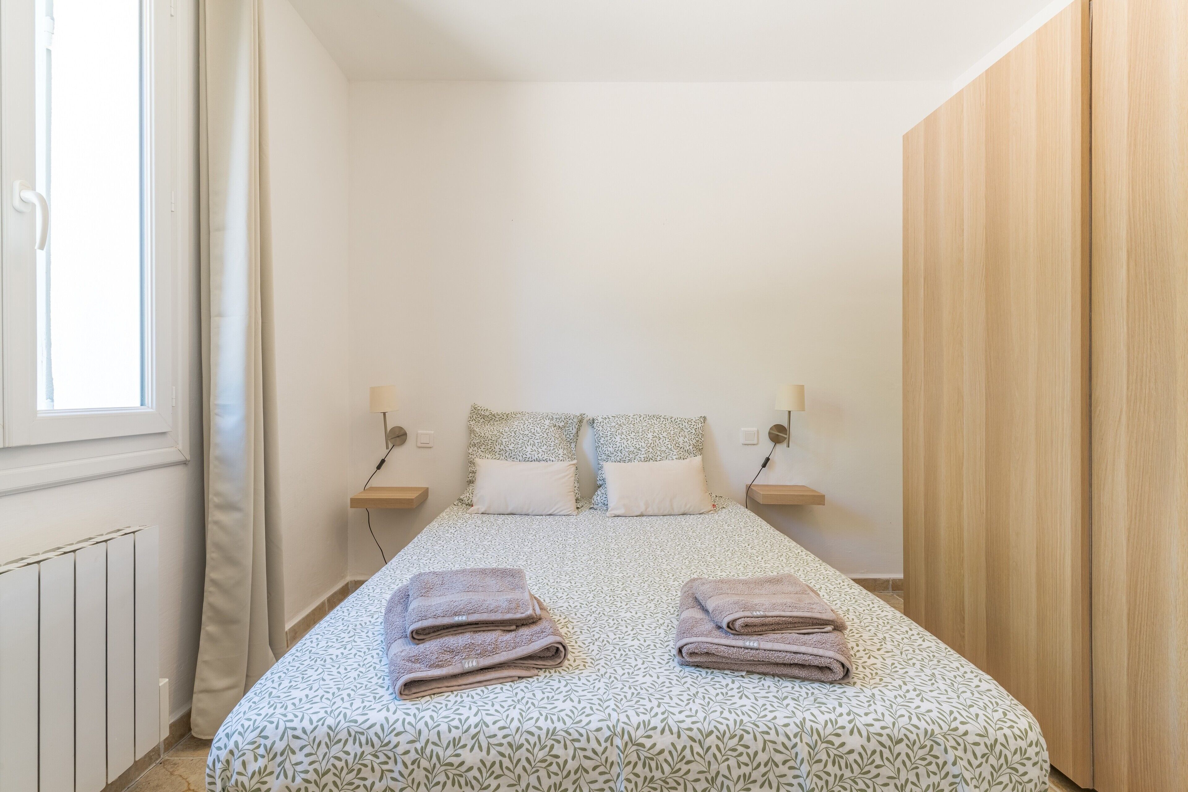 A bedroom with a bed, two pillows, a nightstand, a wooden wardrobe, and a window with curtains.