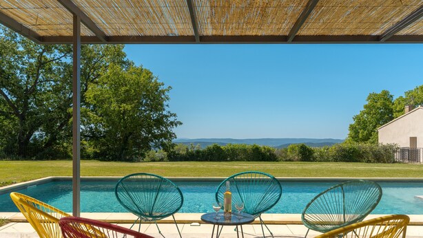 Zona junto a la piscina con sillas y una mesa, vista despejada de la piscina y un paisaje con césped, árboles y colinas al fondo.