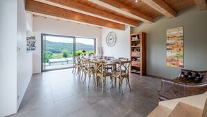 A spacious dining area with a large table and chairs, a wooden ceiling, and a view of greenery outside.