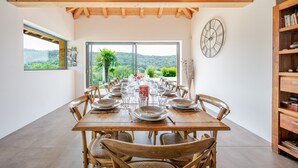 A dining room with a wooden table set for a meal, large windows with a view of greenery, and a wooden ceiling.