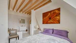 A bedroom with a bed, a chair, a vanity, and a painting on the wall.