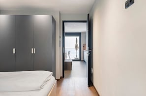 Un pasillo estrecho que conduce a un balcón con vistas, una cama con ropa blanca y un armario de color oscuro.