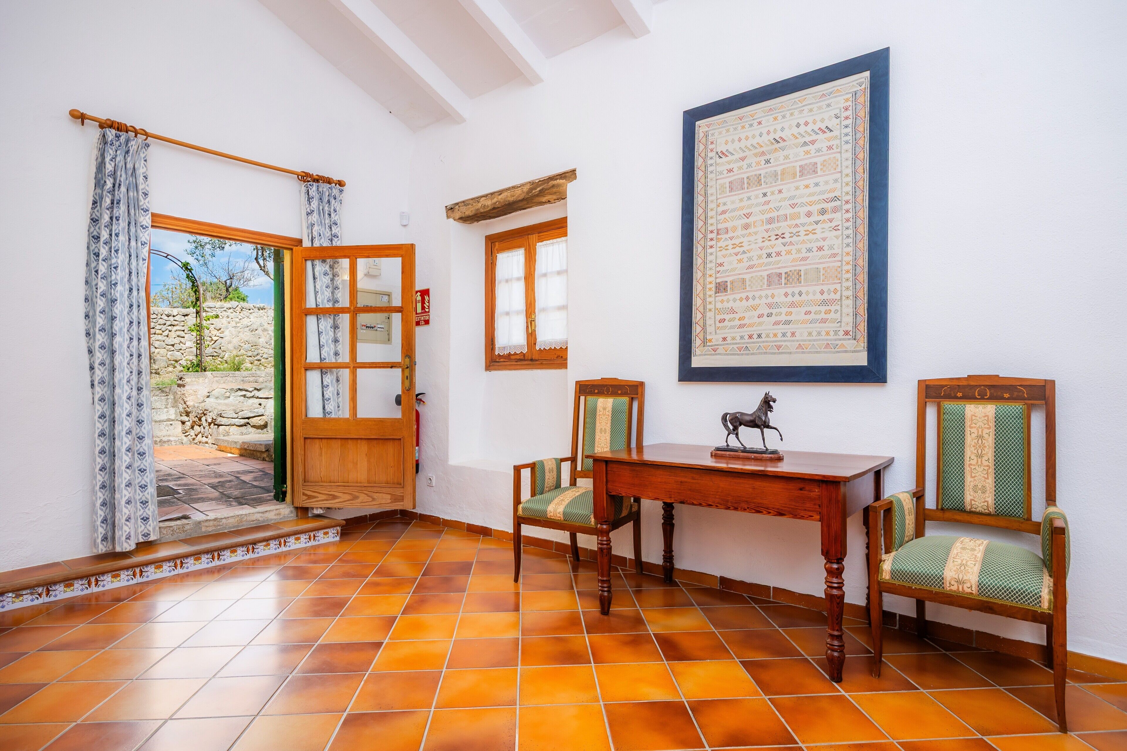A room with a wooden table, two chairs, a framed tapestry, and a door leading outside.