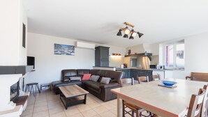 A modern living room with a sofa, coffee table, and dining table. There is a kitchen area with cabinets and a stove. - Gîte d'Ayzi – Charming Rental with Panoramic Views, Ideal for Families or Friends, 15 Minutes from Lourdes (Ouzous)
