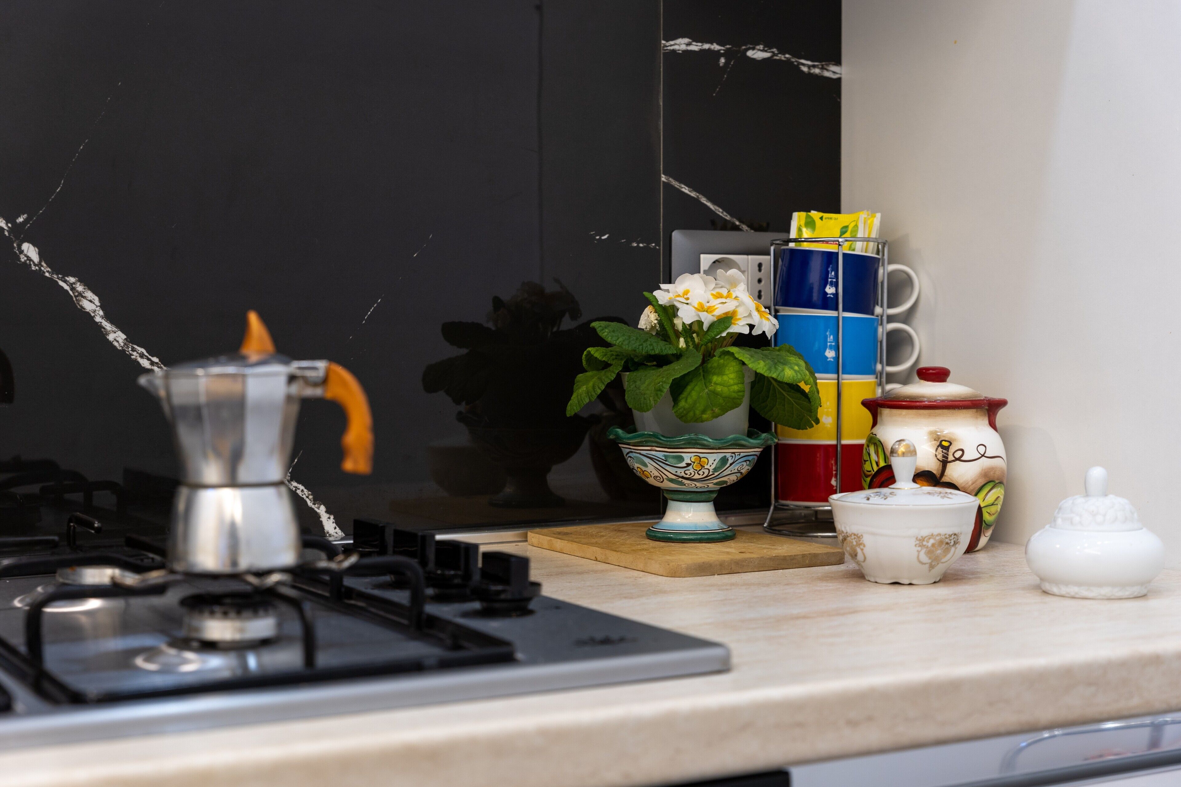 Eine Küche mit einem Gaskochfeld, einer Kaffeemaschine, einem Blumentopf, einer Stapel bunter Tassen und einem dekorativen Keramiktopf.