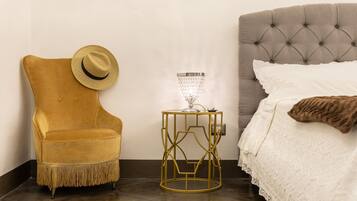 Una habitación con una cama de cabecera capitonada, una mesita de noche con marco dorado y una lámpara, un sillón amarillo mostaza con un sombrero y una colcha blanca.