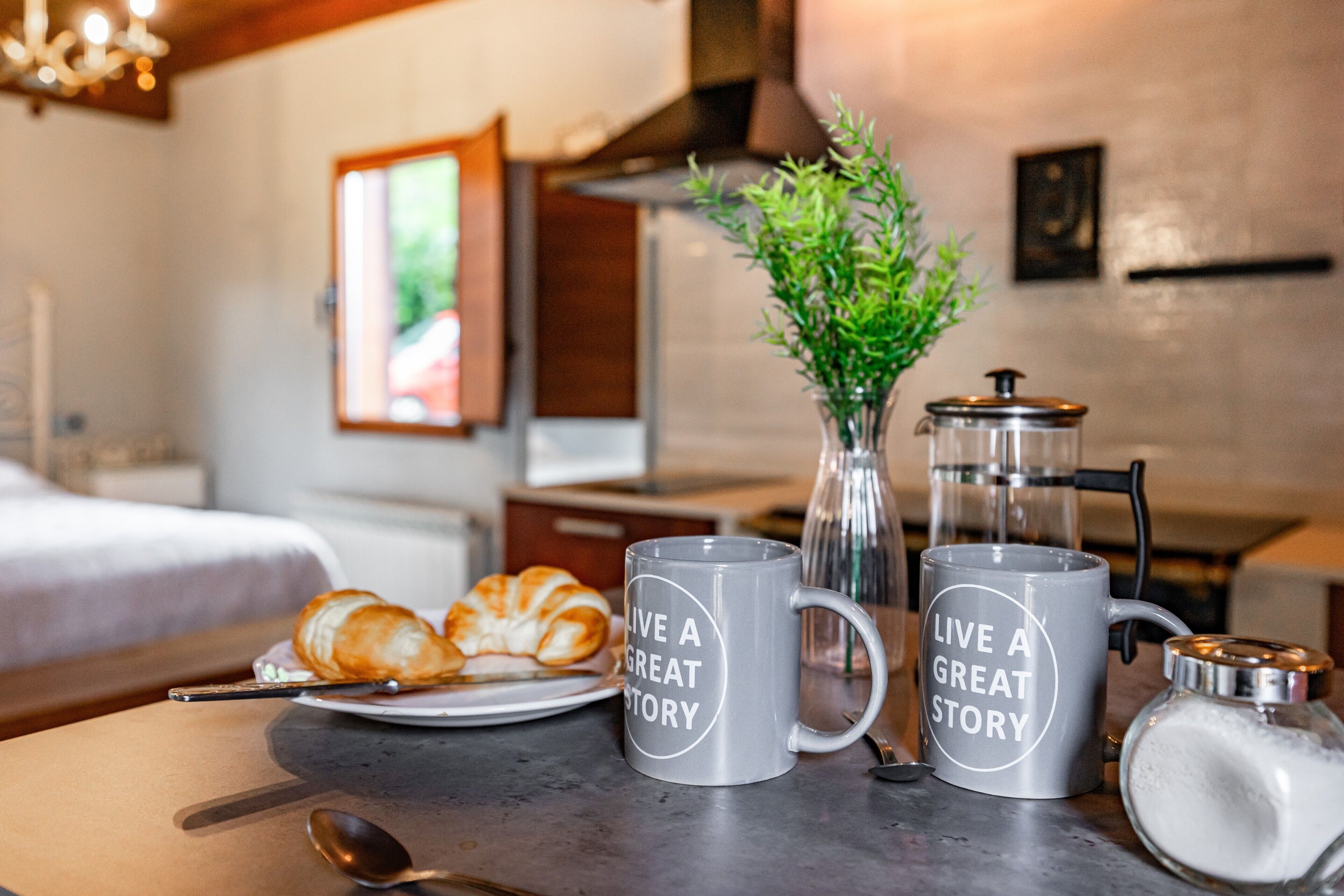Una mesa con dos tazas, un croissant, una cuchara, un vaso de agua y una cafetera.