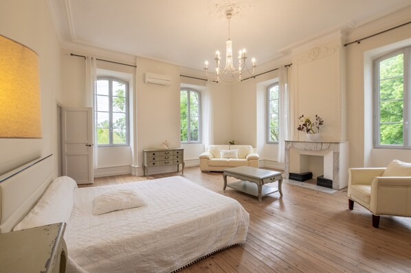 Une chambre spacieuse avec un grand lit, un coin salon doté d’une cheminée et de nombreuses fenêtres laissant entrer la lumière naturelle.