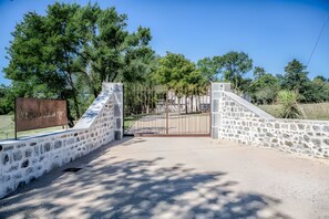 A gated entrance with stone walls and a metal gate, surrounded by trees and a clear sky.