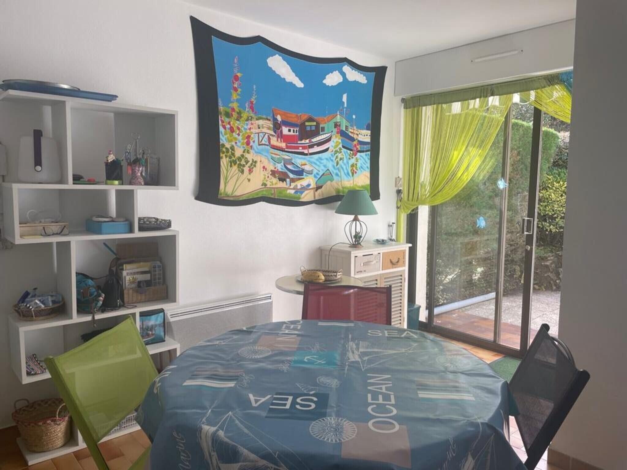 Een kamer met een eettafel, bedekt met een blauwe tafelkleed, een groot schilderij van een kustlandschap en uitzicht op groen via een schuifdeur.