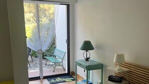 Une chambre à coucher avec un lit, une couvre-lit à motifs verts, une lampe verte, une lampe blanche, une petite table avec une lampe, et une vue sur un patio avec une chaise et un hamac.