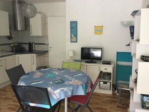 Um espaço compacto com cozinha compacta, área de refeições com mesa e cadeiras, televisão e uma pequena estante com objetos.