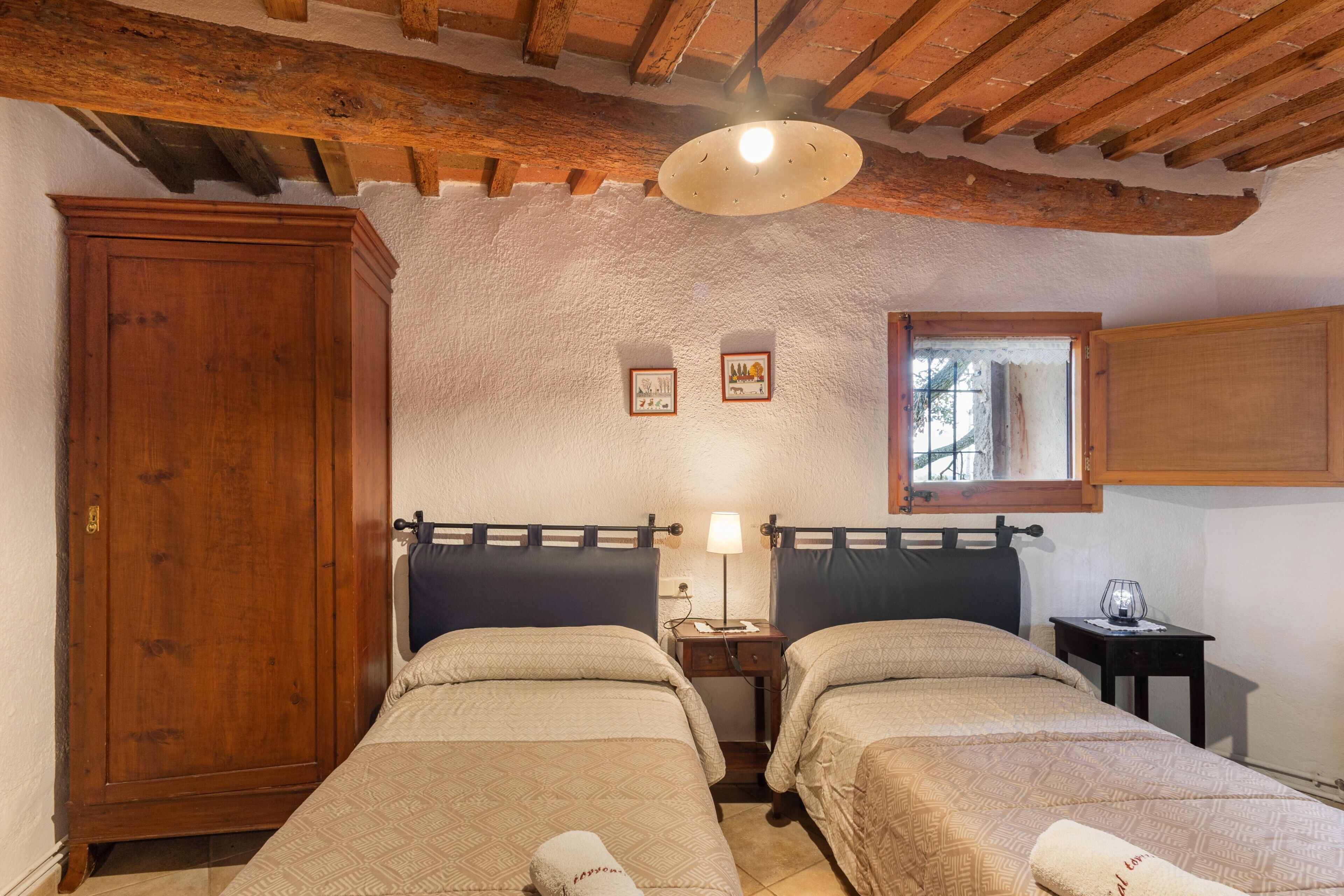Habitación con dos camas, vigas de madera en el techo, un armario y una pequeña mesa con una lámpara.
