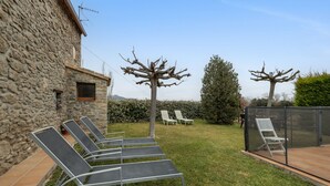 A stone building with a garden featuring lounge chairs and a fenced area.