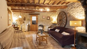 A rustic interior with exposed wooden beams, a stone wall, a large wooden wheel, a sofa, a table, and a fireplace.