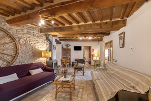 A rustic interior with exposed wooden beams, stone walls, a large round window, and a cozy seating area with a sofa and armchairs. - Country House 'Cal Torrentí' with Mountain View, Private Pool and Wi-Fi (Casserres)