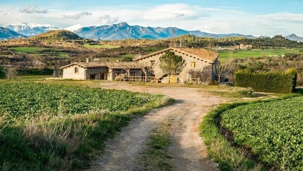 A rustic stone building with a tiled roof, surrounded by green fields and mountains in the background.