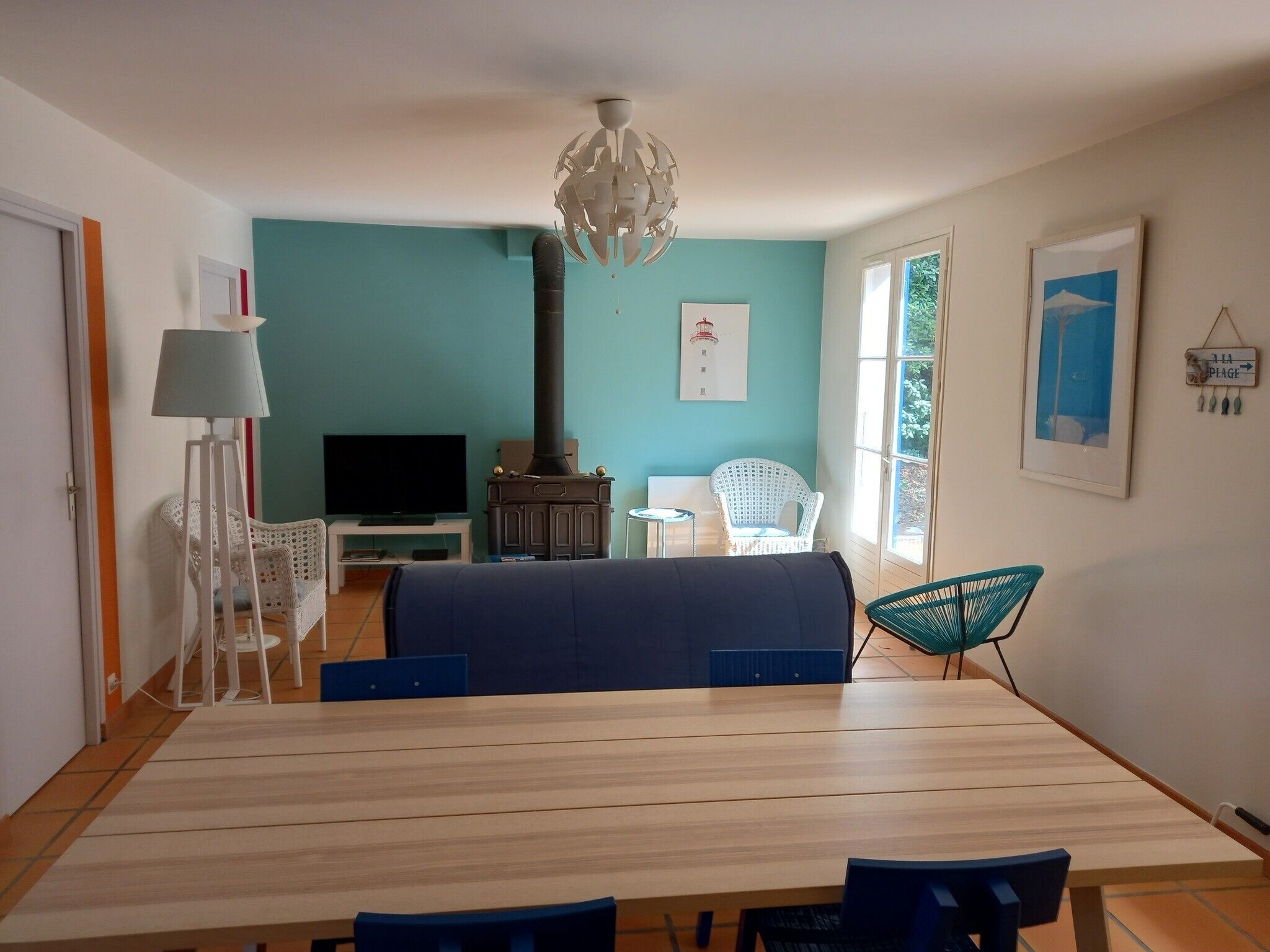 Een kamer met een houten eettafel, een televisie, een witte stoel, een blauwe bank, een witte vloerlamp, een houten kast en een groot raam.