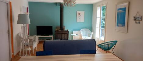Una habitación con una mesa de comedor de madera, un televisor, una silla blanca, un sofá azul, una lámpara de pie blanca, un mueble de madera y un ventanal grande.