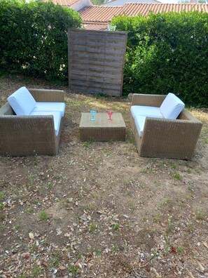 Mobiliario de mimbre exterior con cojines blancos, una mesa cuadrada y un fondo de valla de madera y arbustos verdes.