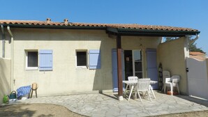 A house with a tiled roof, two windows with shutters, a patio with stone flooring, and outdoor furniture. - Tranquil Escape: House with Private Garden, 150m from the Forest and 20min to the Beach (Saint-Trojan-les-Bains)