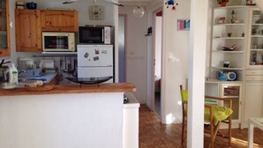 A kitchen with wooden cabinets, a microwave, a refrigerator, and a dining area with a chair and table. - Tranquil Escape: House with Private Garden, 150m from the Forest and 20min to the Beach (Saint-Trojan-les-Bains)