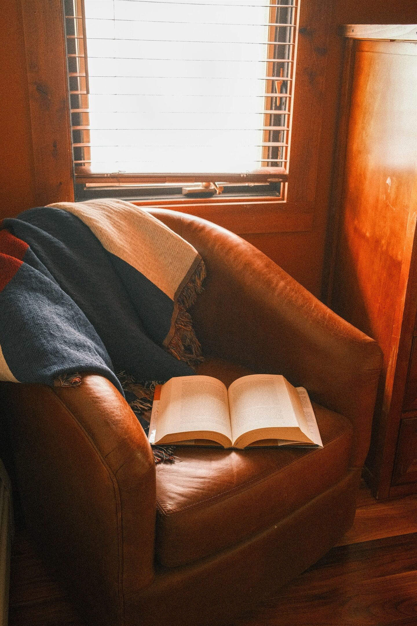 A leather armchair with an open book, positioned near a window with blinds.