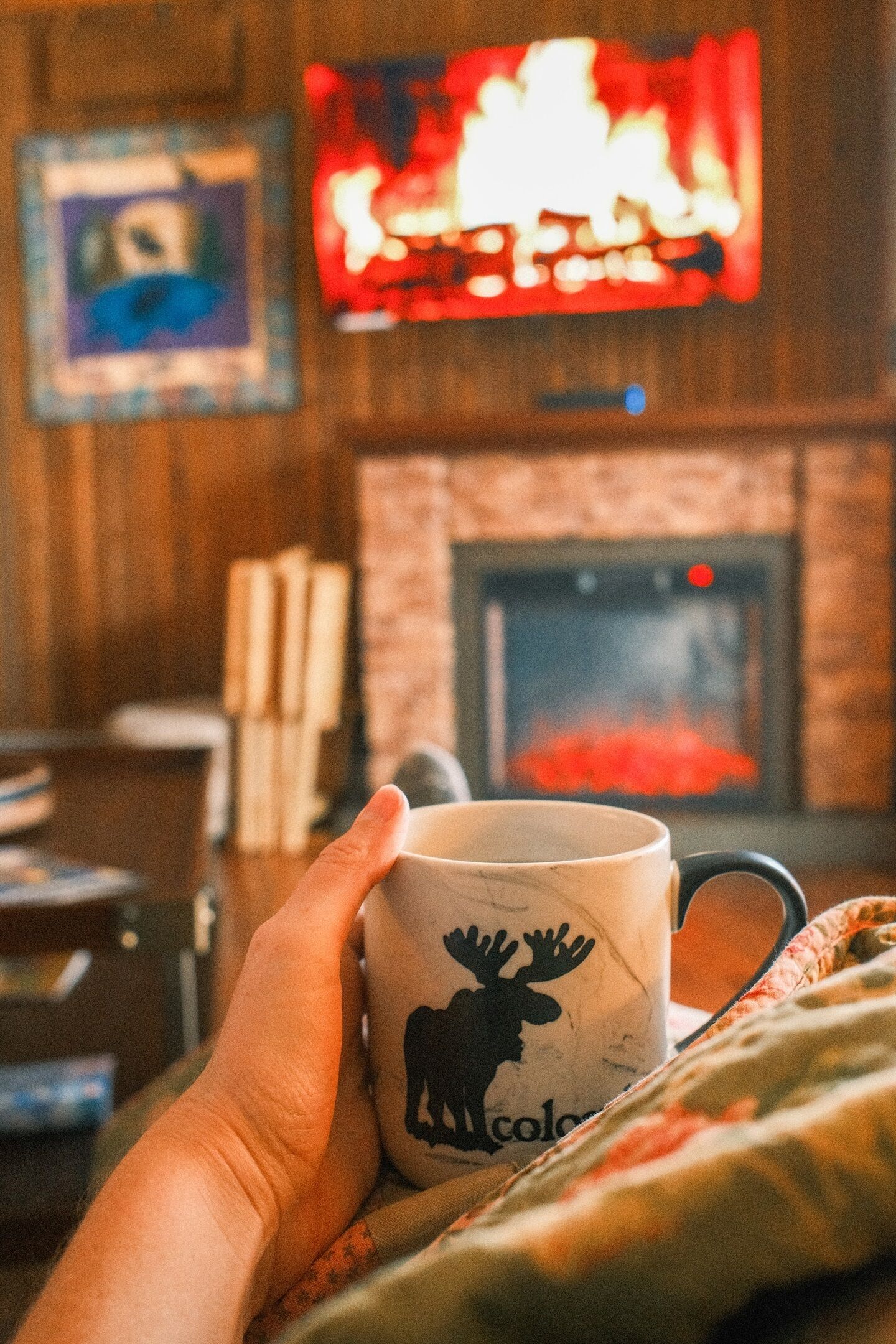 A hand holding a mug with a moose design, a fireplace, and a television displaying a fire.