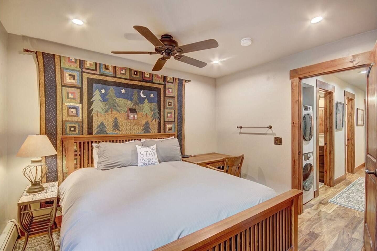 A bedroom with a bed, a nightstand, a lamp, a ceiling fan, a wooden headboard, a wall tapestry, and a wooden door leading to another room.