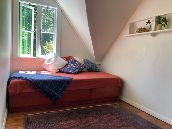 Un dormitorio pequeño y acogedor con una cama roja, una ventana con follaje verde en el exterior, una estantería con objetos decorativos y una alfombra estampada en el suelo.