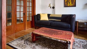 Una sala de estar con un sofá oscuro, un tapete estampado, una mesa de centro de madera y una puerta con paneles de vidrio.