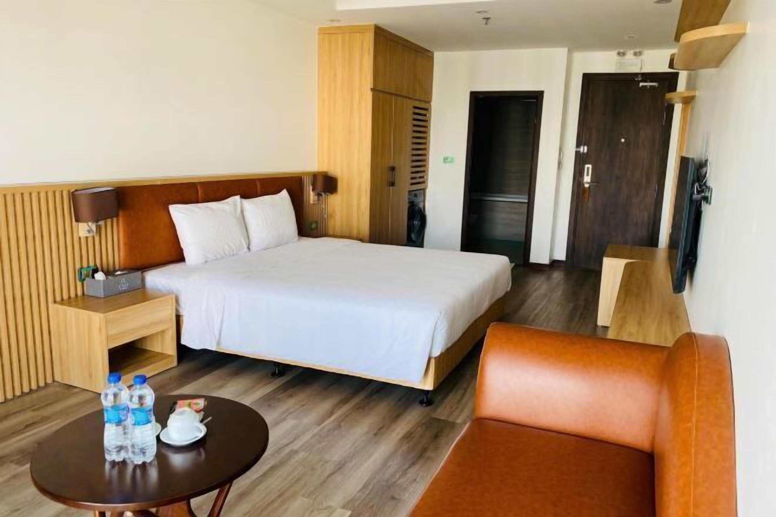 A hotel room with a bed, a wooden wardrobe, a small table with water bottles, a sofa, and a television.