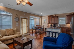 A spacious living room with a sofa, coffee table, and blue armchair. A dining area with a table and chairs is adjacent to a kitchen with wooden cabinets and a refrigerator.